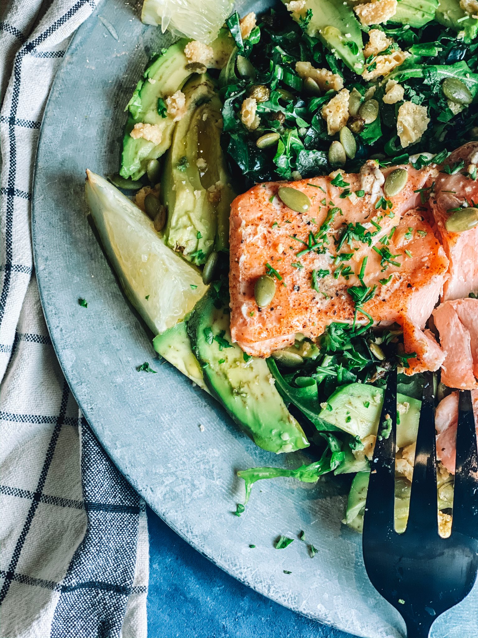 Salmon filet on a bed of greens and avocado, plated with a fork and a blue background.