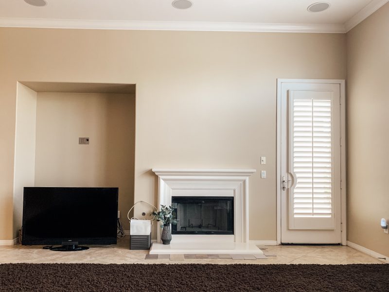 View of the cream colored living room wall with an empty nook, tv sitting on the floor, white fireplace and mantle, and white door leading to the backyard.