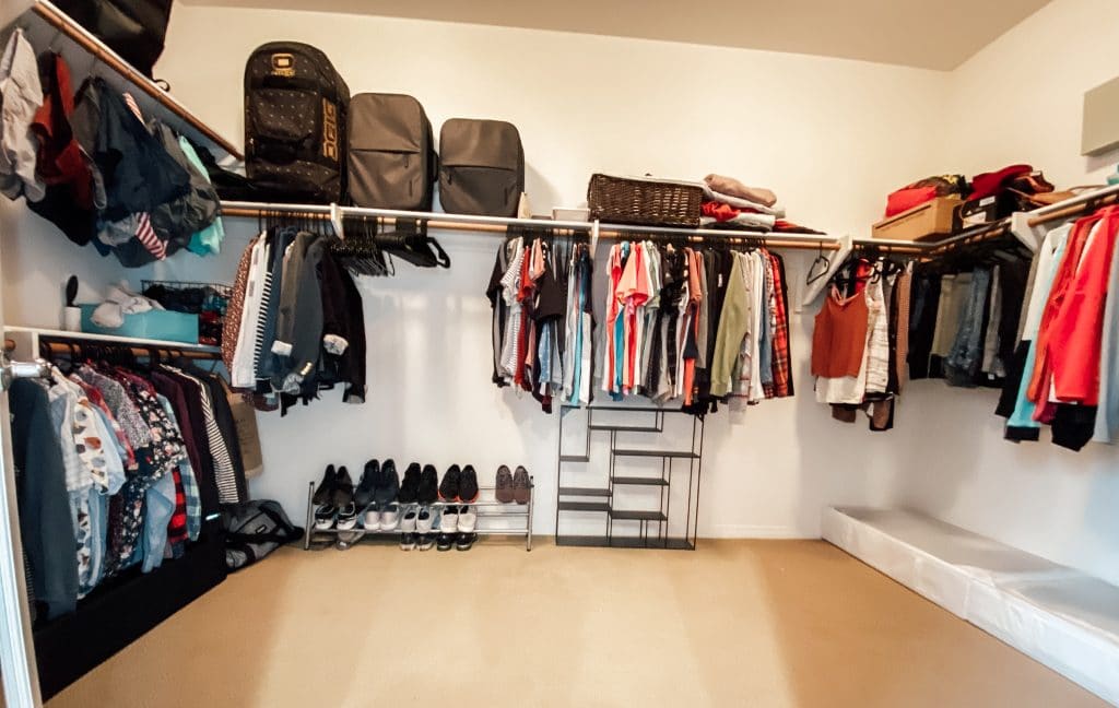 closet with clothes hanging on rods and luggage on shelf above